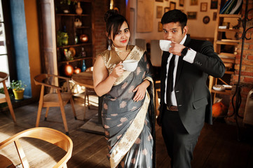 Elegant and fashionable indian friends couple of woman in saree and man in suit sitting on cafe and drinking tea.