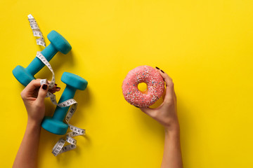 Female hand holding sweet donut, measuring tape, dumbbells over yellow background. Top view, flat...