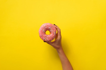 Woman hand holding delicious pink donut on yellow color background. Top view banner with copy...