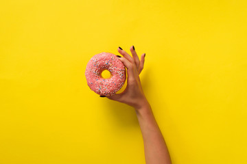Female hand holding pink donut over yellow background. Top view, flat lay. Sweet, dessert, diet...