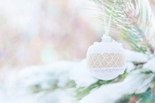 Christmas winter holiday greeting card.  White rustic Christmas ornament ball with burlap on green christmas trees with snow. Festive outdoor background. Xmas composition decoration