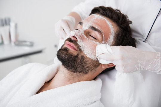 Relaxed man closing eyes and cosmetologist wiping out mask