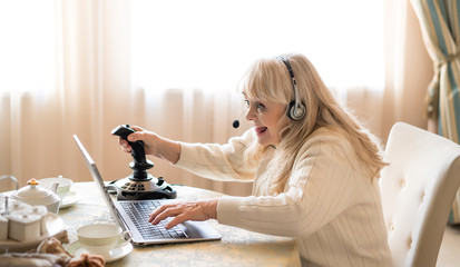 Senior woman plays video games with joystick on a laptop - obrazy, fototapety, plakaty