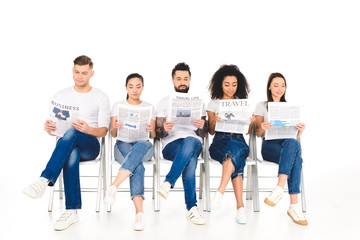 Obraz premium multiethnic group of young people sitting on chairs with crossed legs and reading newspapers isolated on white