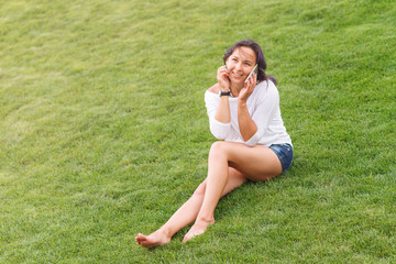 Fototapeta premium Happy beautiful young woman sitting on grass and using smartphone
