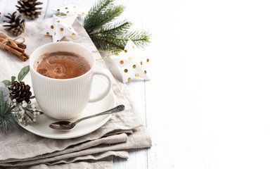 Cup of hot chocolate with cinnamon sticks on the white wooden table. Winter cocoa drink on a napkin, Christmas-tree golden stars decorations, branches and cones
