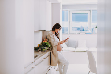 Progressive pregnant mum using tablet and standing on kitchen