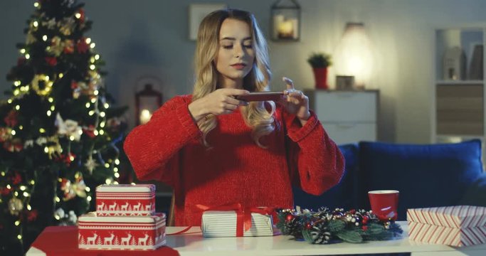 Caucasian young attractive woman taking photo of the flat lay Christmas present with decorations in her living room with a beautiful x-mas tree.