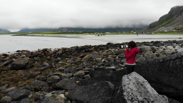 Tourist woman on rocky seashore taking photo with camera from sea coast at cloudy hazy day. Skagsanden Beach Flakstadoy Lofoten archipelago Norway