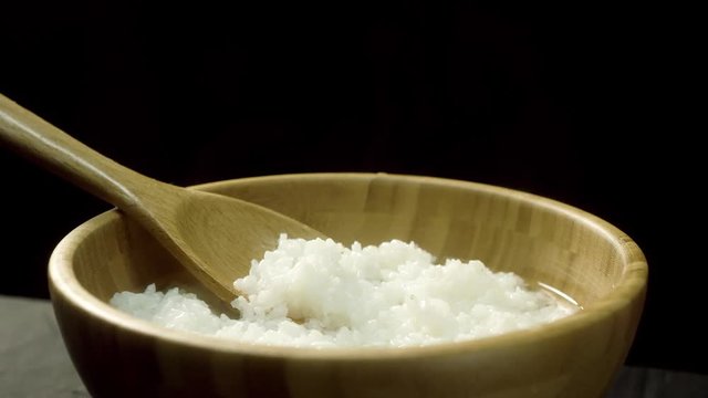 Close up for steamed rice in a wooden bowl with wooden stirring spoon, isolated on black background. Freshly cooked rice with steam in deep, brown plate with big spoon, food, cooking concept.