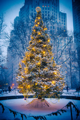 Snow-covered Christmas tree with golden lights glowing against a stark urban background after a...