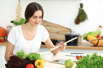 Beautiful Hispanic woman cooking in kitchen while using tablet computer and wooden spoon. Housewife found new recipe for dinner or breakfast. Healthy meal and householding concepts