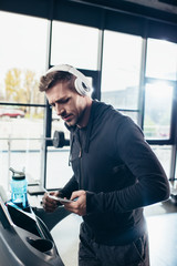 Naklejka na ściany i meble handsome sportsman in hoodie exercising on treadmill and using smartphone in gym