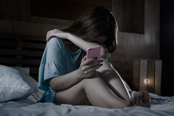 young depressed Asian Chinese girl using mobile phone crying on bed at night feeling sad and...