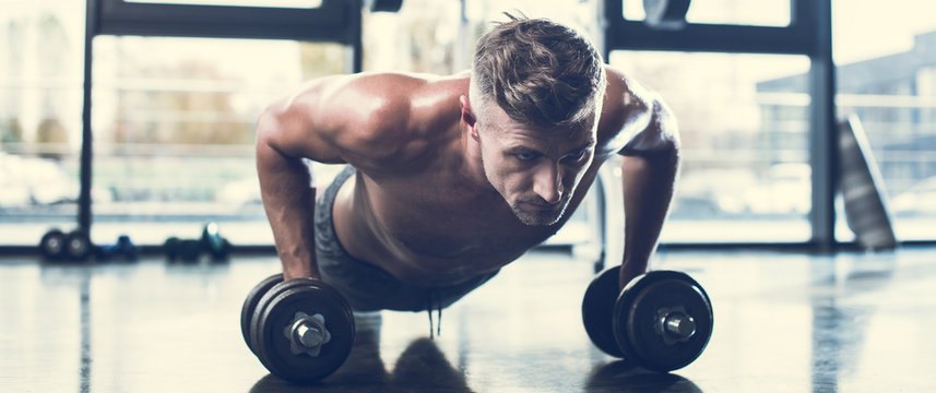 handsome shirtless sportsman training with dumbbells and doing plank in gym