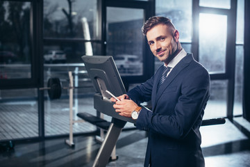 handsome businessman in suit leaning on treadmill and using smartphone in gym