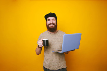 Cheerful bearded man looking at the camera holding a cup of coffee or tea and holding laptop