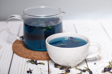 Obraz na płótnie Canvas Butterfly pea blue flower tea. Healthy detox herbal drink. Butterfly pea blue tea in white Cup and teapot top view on white wooden table with scattered dried flowers