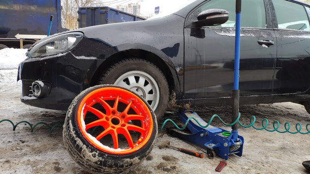 Winter tire fitting on the street. Wheel service, Tire repair, and changing summer to winter tires. Black car lifted on jack. Summer tires in winter. Orange rims