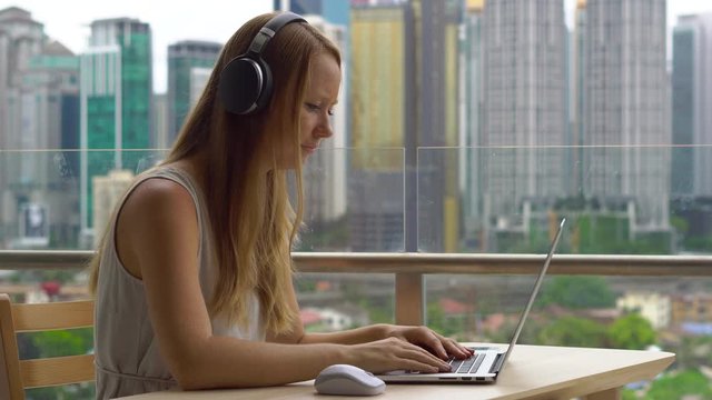 Young woman freelancer workes on her laptop at a balcony with a background of a city center full of skyscrapers. Remote techer has a conversation with a student through the internet
