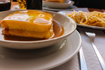 Traditional portuguese sandwich francesinha with glass of wine and fried potato. Francesinha in hot sauce with fork and knife. Portuguese cuisine and food concept. Meat and cheese meal. Lisbon lunch.