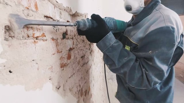 worker in protective suit demolishes plaster wall. Dirty, hard work. Personal protective equipment. Helmet, respirator and goggles.