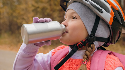 Fototapeta premium Kid drink water from an aluminum flask bottle. One caucasian children rides bike road in autumn park. Little girl riding black orange mtb cycle in forest. Biker motion ride with backpack and helmet.