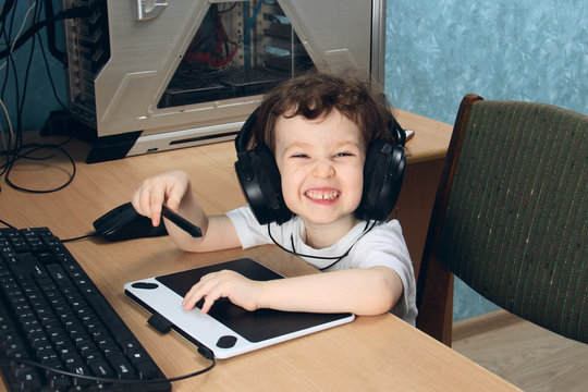 Little 2 3 year old baby girl in white clothers draws at the home computer in graphics drawing tablet. The child is holding a pen and laughs. On the head are huge headphones. close up