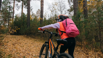 Fototapeta premium One caucasian children walk with bike in autumn park. Little girl walking black orange cycle in forest. Kid goes do bicycle sports. Biker motion ride with backpack and helmet. Mountain bike hardtail.