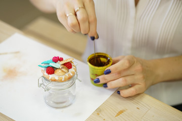 woman sitting at a white table and creates a polymer clay kitchen jar with decorations in the form of leaves and cookies, step by step master class