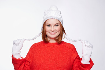 Portrait of cheerful European freckled woman, wearing white knitted mittens and hat with two plaits, looking at camera with cutest lovely smile, dressed in red oversized stylish sweater.