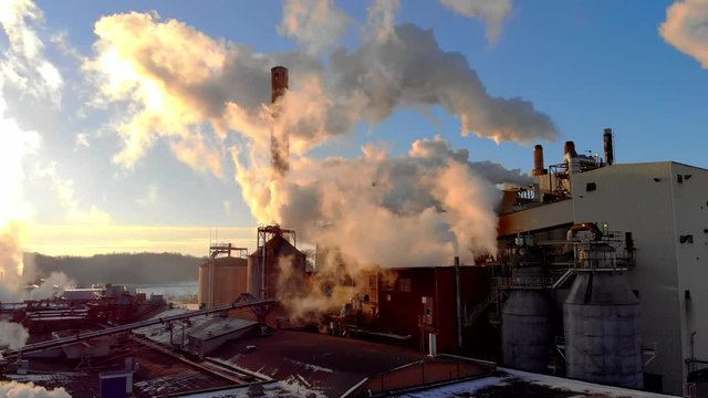 Factory smokestacks, steaming and smoking, stunningly back-lit by the sunrise, moving aerial view.