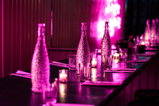 Pink Neon Lights Shining On A Long Wooden Table and Stools With Glass Bottles, Glasses, Napkins and Flatware.
