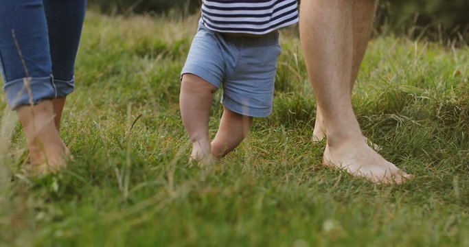 Close up of the first steps of the baby while holding hands of mother and father from bothe sides on the green grass. Caucasian barefeet. Outside.