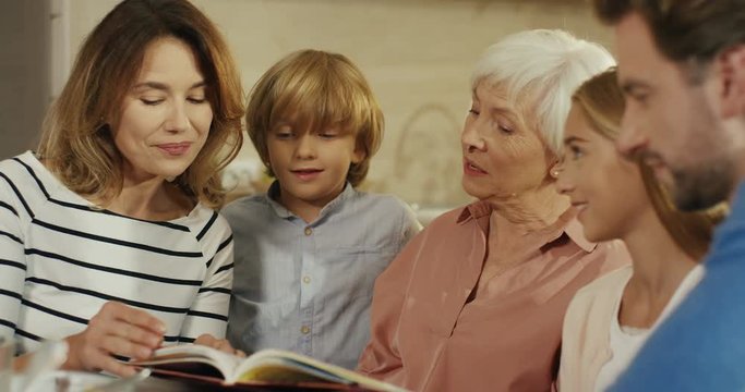 Caucasian senior woman with daughter or son and grandchildren looking at some book while reading and flipping pages at home. Close up. Indoors.