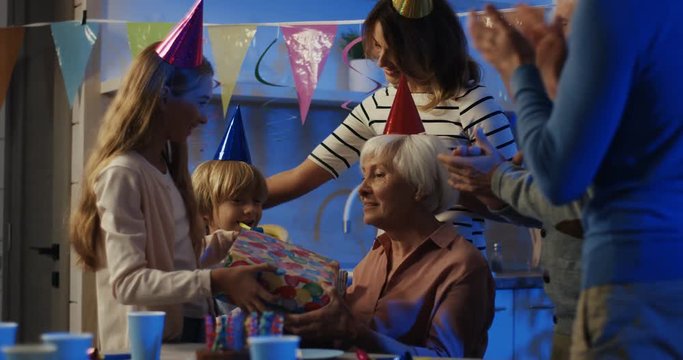 Little teen cute cheerful grandchildren wishing health to their grandmother, kissing her and giving present on her birthday. Indoors.