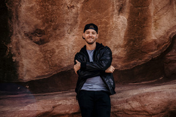 Portrait of Young Good Looking Casual Traveling Handsome Man Smiling Near Ancient Desert Red Rocks in Jacket Outside