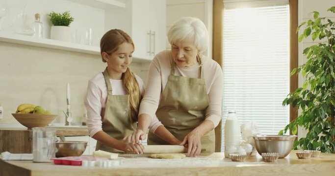Nice grey haired grandmother teaching her little teenage granddaughter making a dough in the modern kitchen.