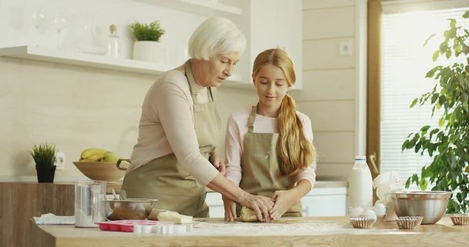 Cute and pretty Caucasian teen girl learning from her grandmother to knead a dough in the cozy kichen. Indoor.