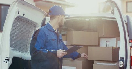 Caucasian mailman in the blue uniform standing at the white van and checking carton parcels, then writing down info. Outdoors in the sunlight.