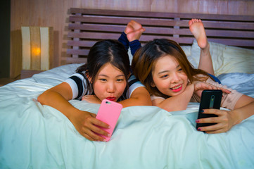 young happy and pretty Asian Korean girlfriends sitting at home bedroom laughing and talking having fun using internet social media app with mobile phone sitting on bed