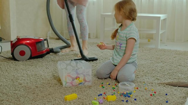 mom and daughter, a young woman cleans up with a vacuum cleaner, a little girl with blond hair collects toys, the designer in a container, helps mom