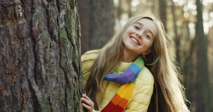 Portrait of the Caucasian blonde pretty cute girl peering from the tree trunk and smiling to the camera in the forest. Close up. Outside.