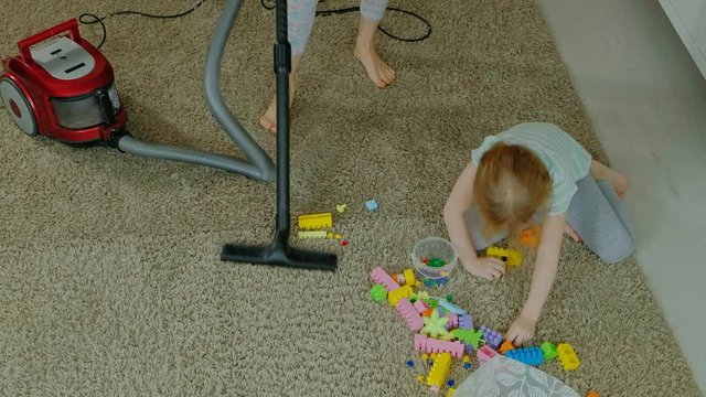 mom and daughter, a young woman cleans up with a vacuum cleaner, a little girl with blond hair collects toys, the designer in a container, helps mom