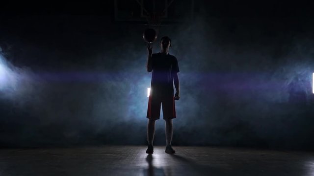 Basketball player performing different tricks and spinning ball in dark gym with smoke. Slow motion