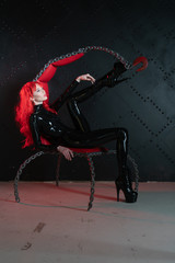 dangerous cruel fetish girl with red hair posing in black bdsm latex clothes at the chair from the...