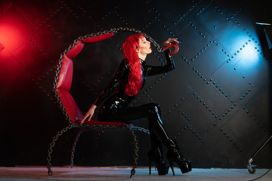 dangerous cruel fetish girl with red hair posing in black bdsm latex clothes at the chair from the chains in the style of sadomasochism