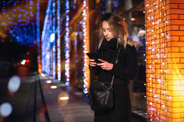Outdoor portrait of young beautiful woman after shopping using her mobile phone near night city