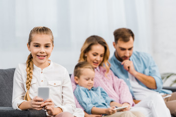 smiling girl sitting on sofa, holding smartphone and looking at camera with family at background in apartment