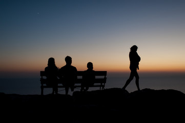 Naklejka na ściany i meble silhouette of a person on a sunset. Three people sitting on a bench and one woman is leaving
